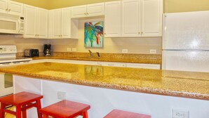 A kitchen with white cabinets, a granite countertop, a microwave, a toaster, a coffee maker, and a refrigerator. There are two red stools at the counter. - Cherry Grove Villas - 204 Villa 6 (North Myrtle Beach)