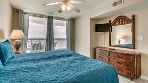 A bedroom with a large bed, a ceiling fan, a dresser, a mirror, and a view of the ocean.