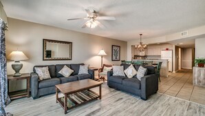 A spacious living room with two sofas, a coffee table, and a ceiling fan. A kitchen is visible in the background.