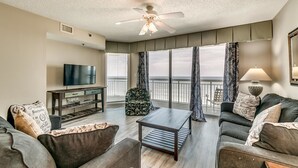 A living room with a flat-screen TV, a ceiling fan, a sofa, a coffee table, and a view of the ocean through large windows.
