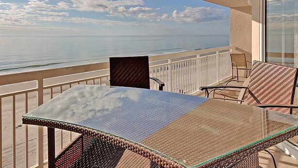 A balcony with a glass table and wicker chairs, overlooking a calm sea and clear sky. - Crescent Shores N - 703 Apartment 4 (North Myrtle Beach)