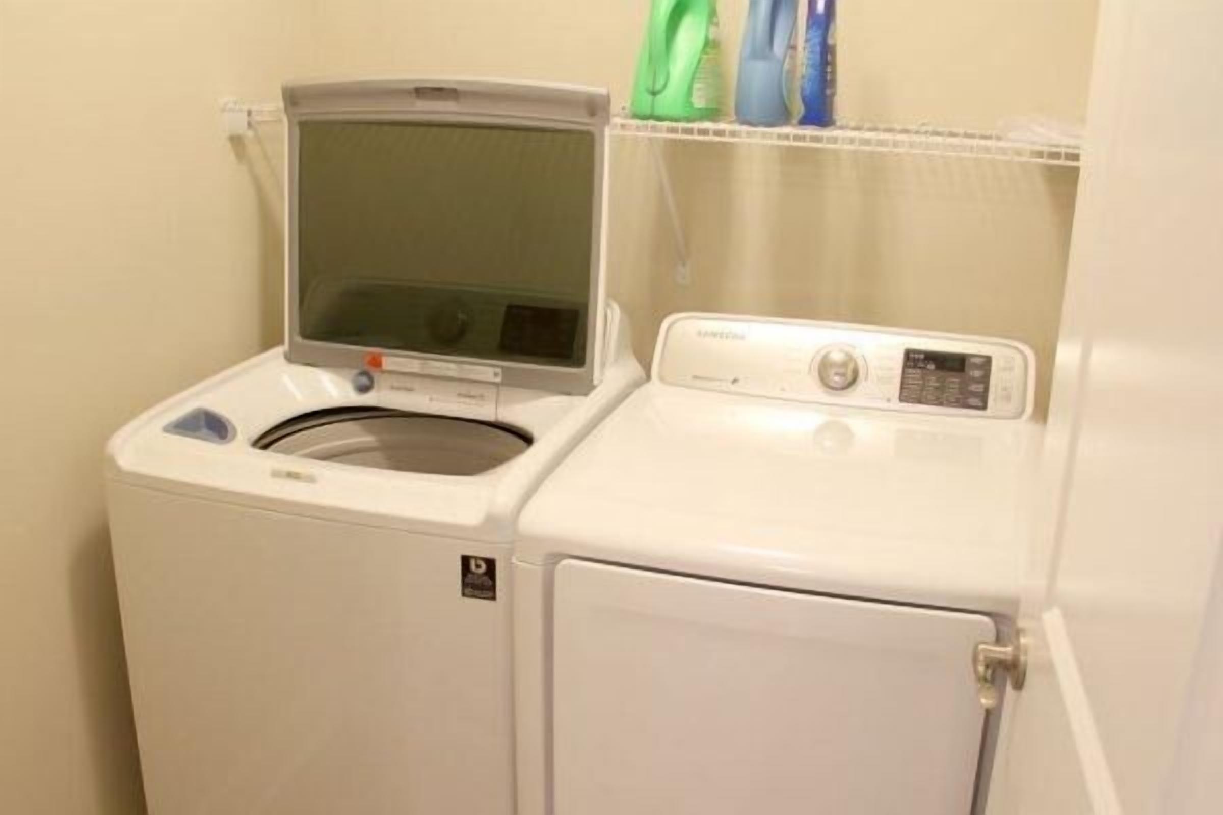 Una lavanderia piccola e compatta, con lavatrice e asciugatrice a carica frontale, un piccolo ripiano con prodotti per la pulizia e una porta con maniglia.