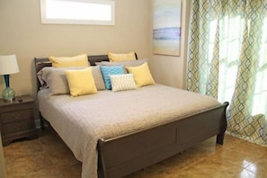 Cama bem arrumada com colcha bege e várias almofadas num quarto com chão de azulejos e cortina estampada.