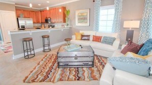 A modern living room with a white sofa, a coffee table with a vintage suitcase, and a kitchen area with wooden cabinets and a stainless steel refrigerator. - South Beach Cottages - 2716 Apartment 3 (Myrtle Beach)