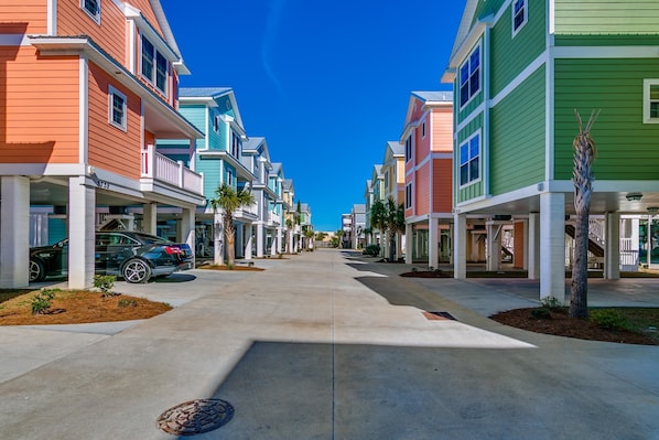 Une rangée de maisons de ville aux couleurs vives, disposées de part et d’autre d’une allée centrale, chacune ayant un garage et un petit jardin devant.