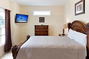 A hotel room with a large bed, a nightstand with a lamp, a dresser, a television mounted on the wall, and a window with curtains. - South Beach Cottages - 2613 Apartment 4 (Myrtle Beach)