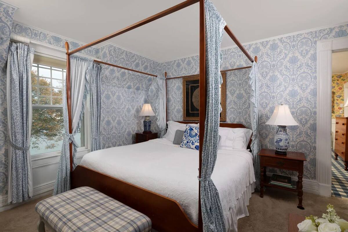 A four-poster canopy bed with a wooden frame, two bedside tables with lamps, a bench, and patterned wallpaper.