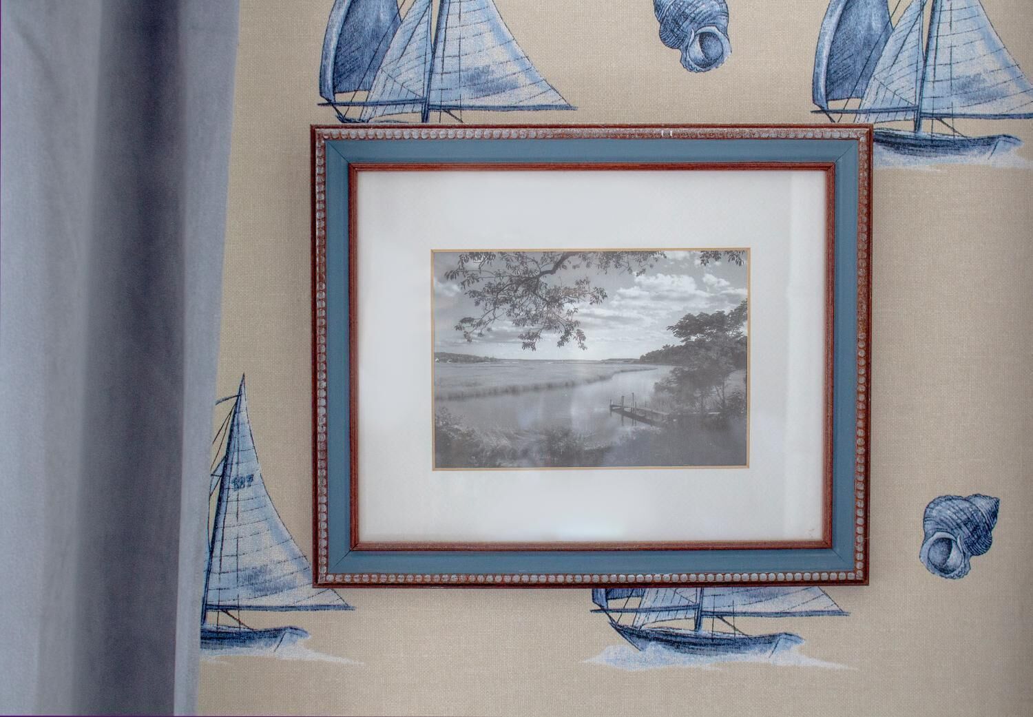 Une photographie encadrée représentant un paysage avec des voiliers et une scène de plage, le tout agrémenté d’illustrations de voiliers sur un mur à texture.
