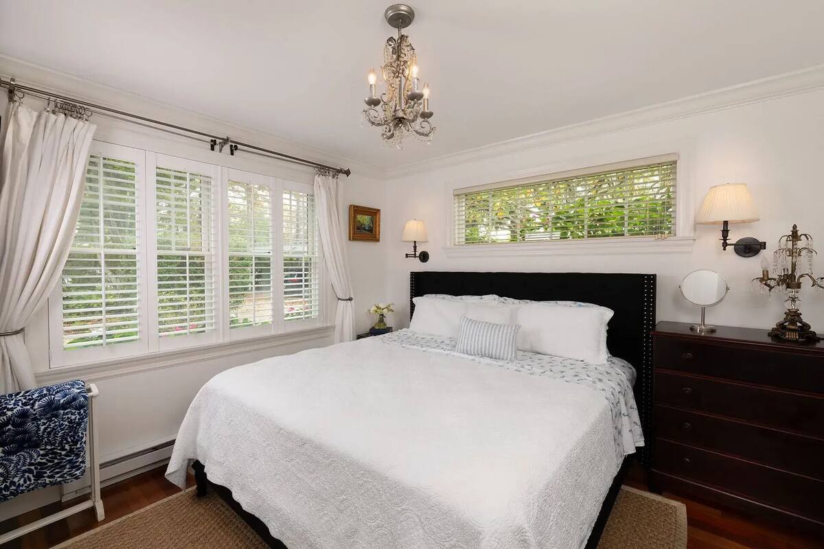 A bedroom with a large bed, a chandelier, a window with blinds, a dresser, and a nightstand.