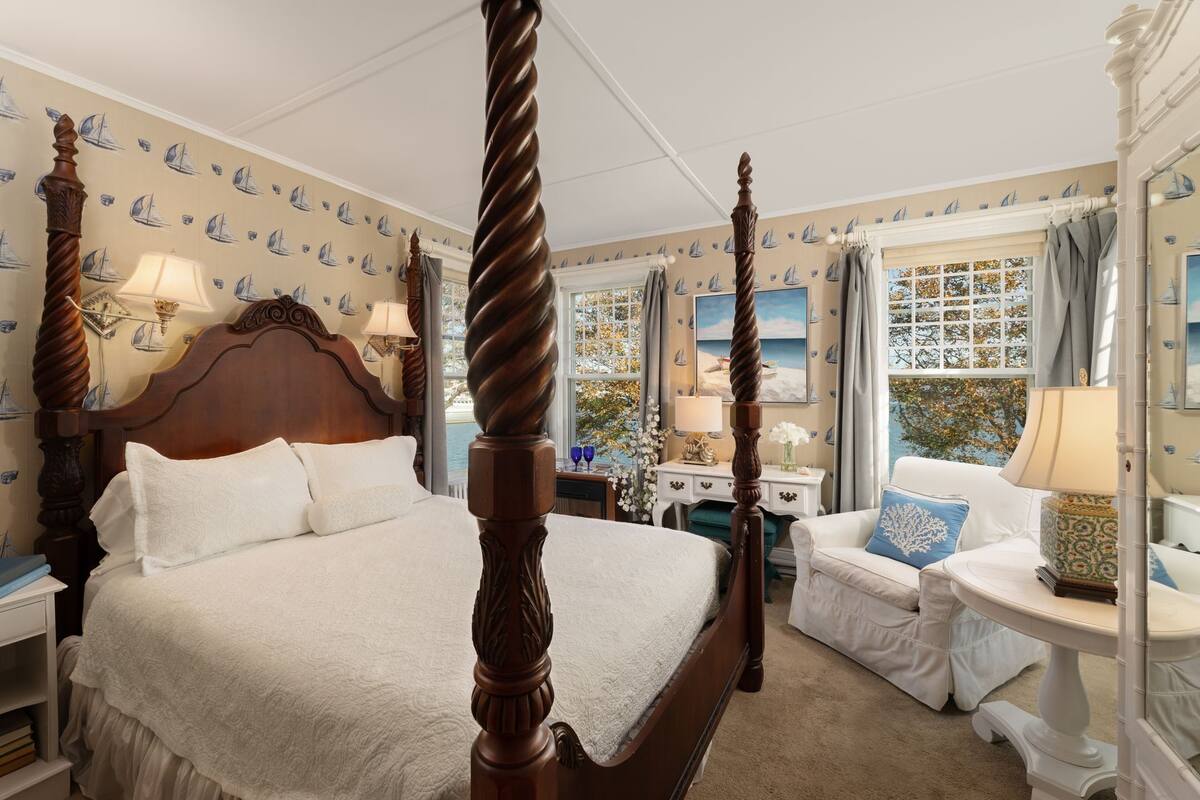 A four-poster bed with a white bedspread, a wooden nightstand with a lamp, a white sofa with a blue pillow, and a window with a view of the sea.