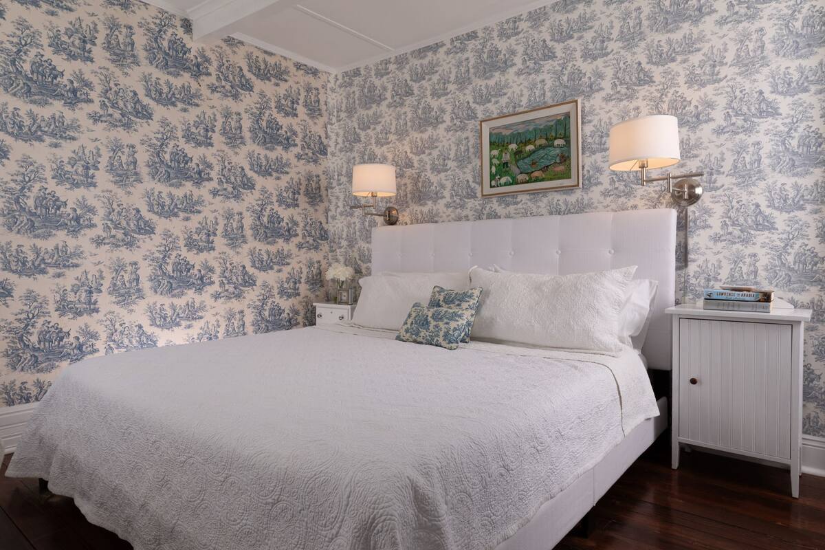 A bedroom with a large bed, white bedding, and a nightstand with a lamp. The walls have a blue floral wallpaper.