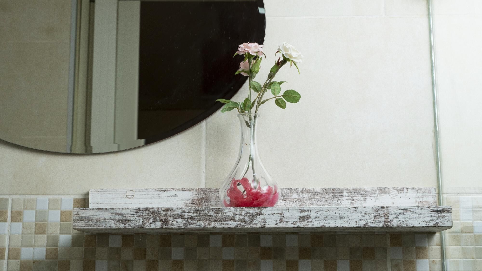 A bathroom with a white tiled wall, a round mirror, and a rustic wooden shelf holding a clear vase with pink and white flowers.
