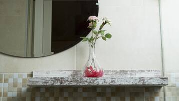A bathroom with a white tiled wall, a round mirror, and a rustic wooden shelf holding a clear vase with pink and white flowers.