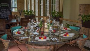 A round glass table set for six with pink cushioned chairs, surrounded by potted plants and a menu board in the background. - Hotel Quinta Moctezuma (Yautepec)