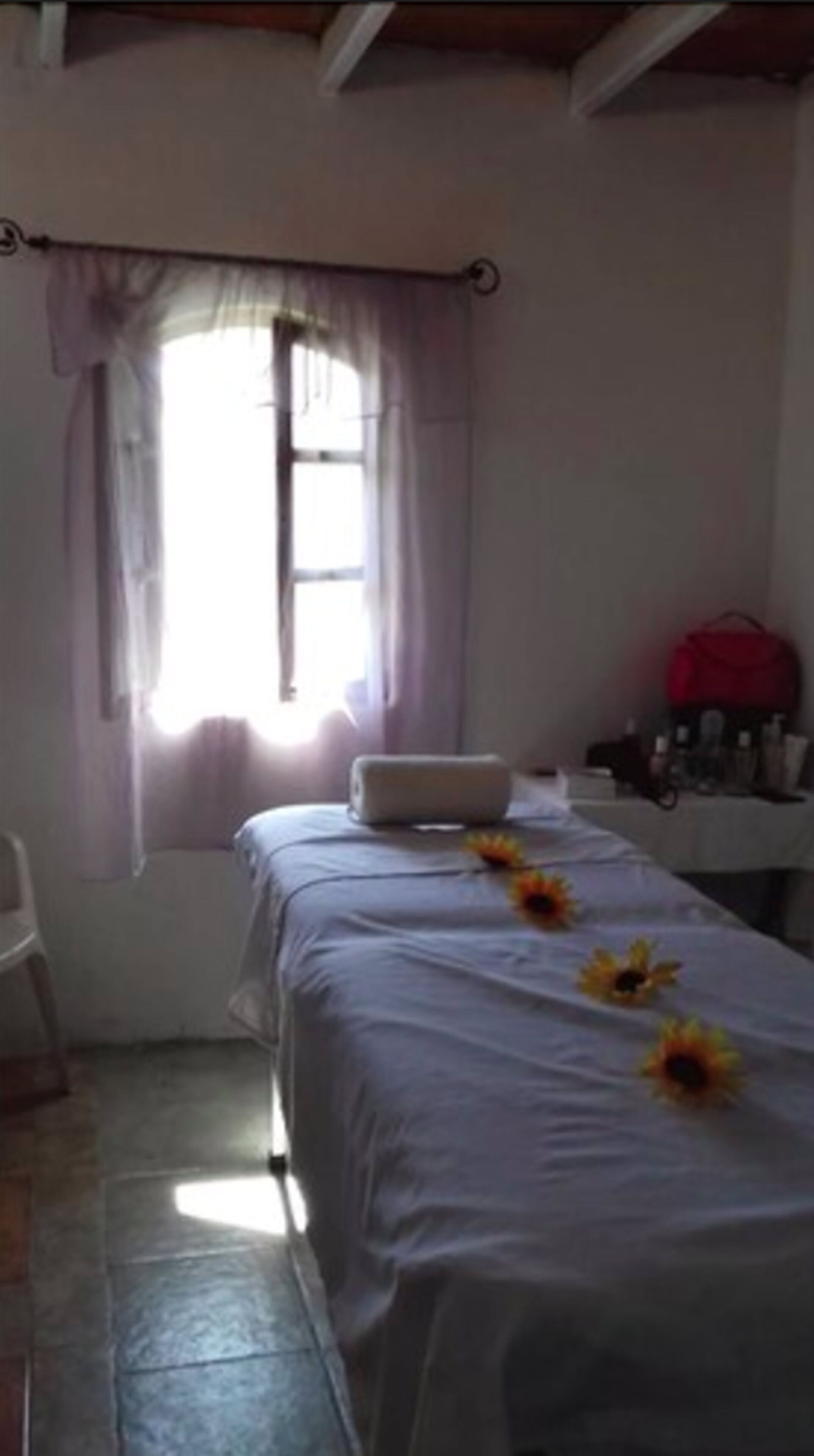 Una habitación con una cama cubierta de sábanas blancas, una ventana con cortinas, una mesita con artículos de aseo y un bolso rojo sobre una silla.