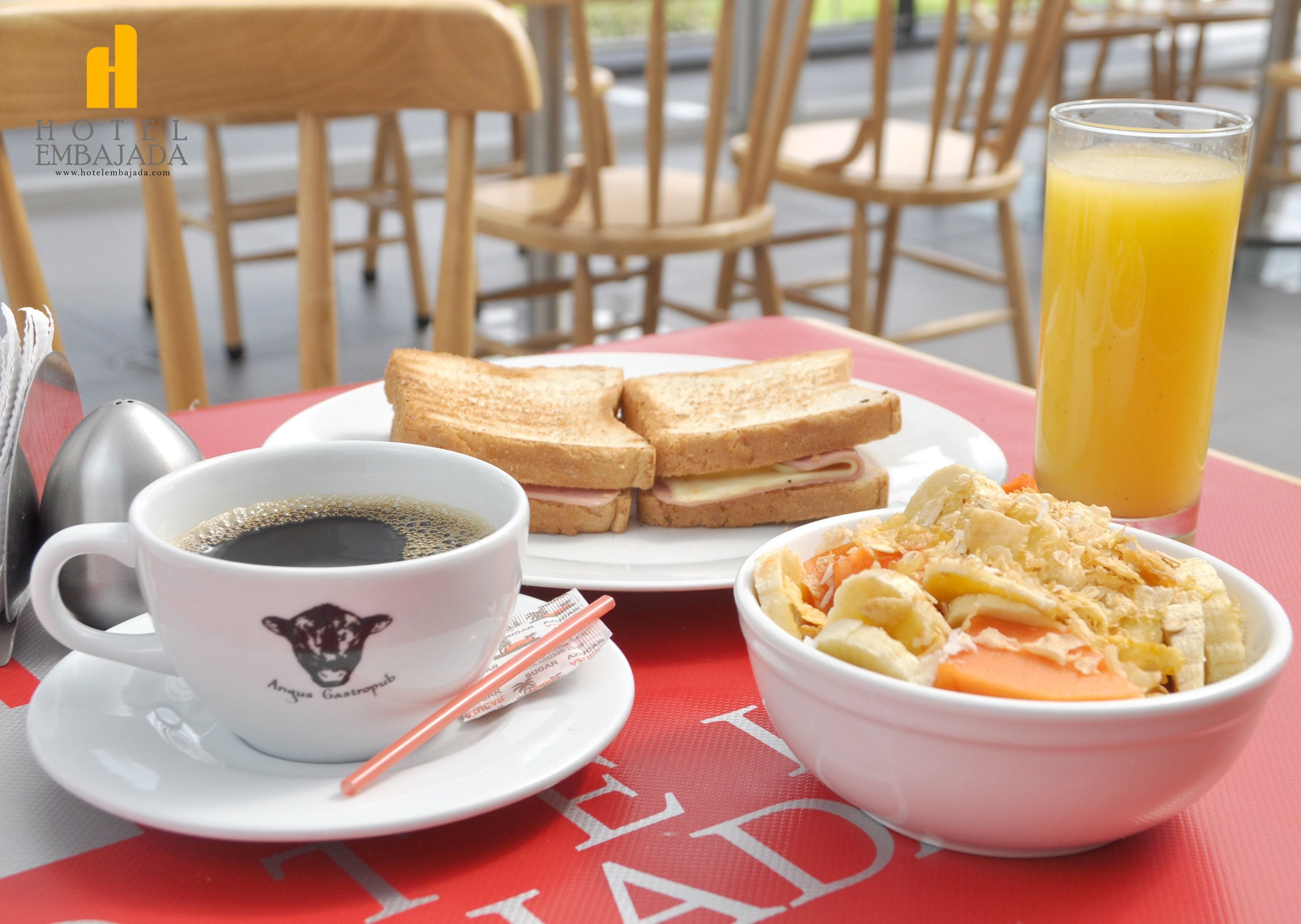 En frukostuppsättning med en kopp kaffe, två skivor rostat bröd med skinka och ost, en skål med flingor och ett glas apelsinjuice på ett rött bord.