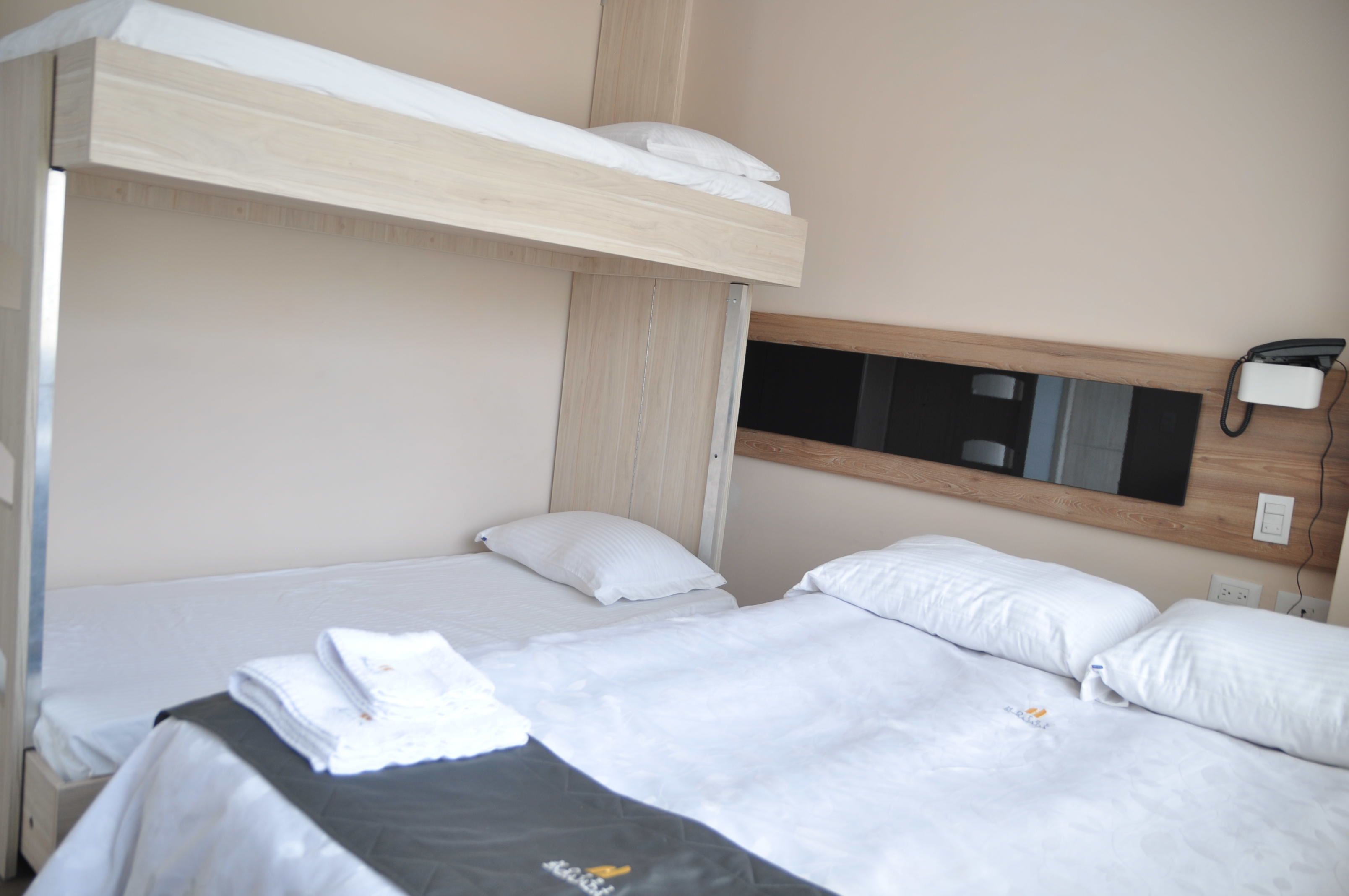 A hotel room with a bunk bed, a flat bed, and a bedside telephone.