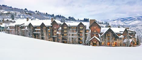 A large, multi-story lodge with a symmetrical facade, featuring a mix of stone and wood construction, set against a snowy mountain backdrop.
