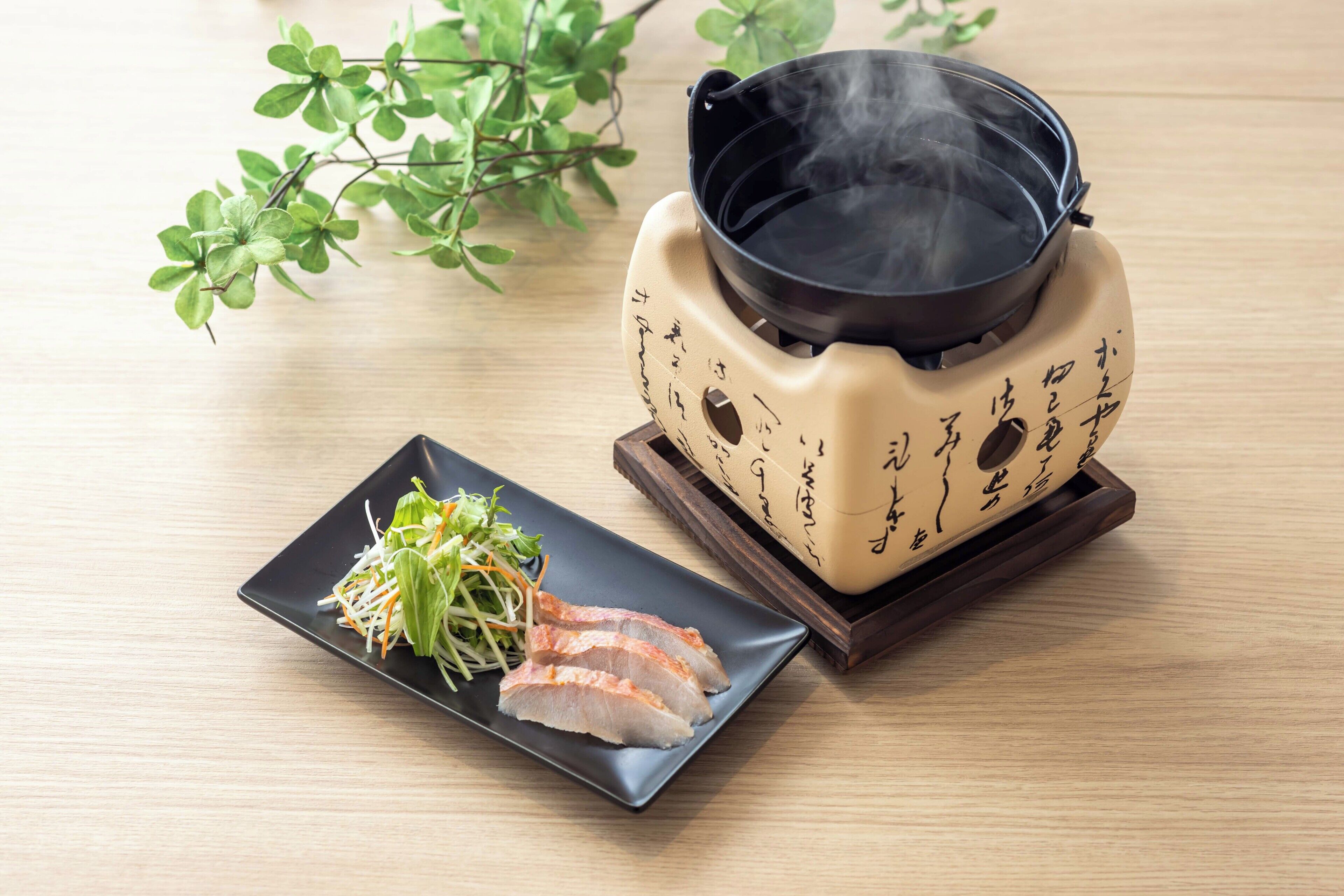 A table with a traditional Japanese hot pot, a plate with sliced meat and vegetables, and a small plant.