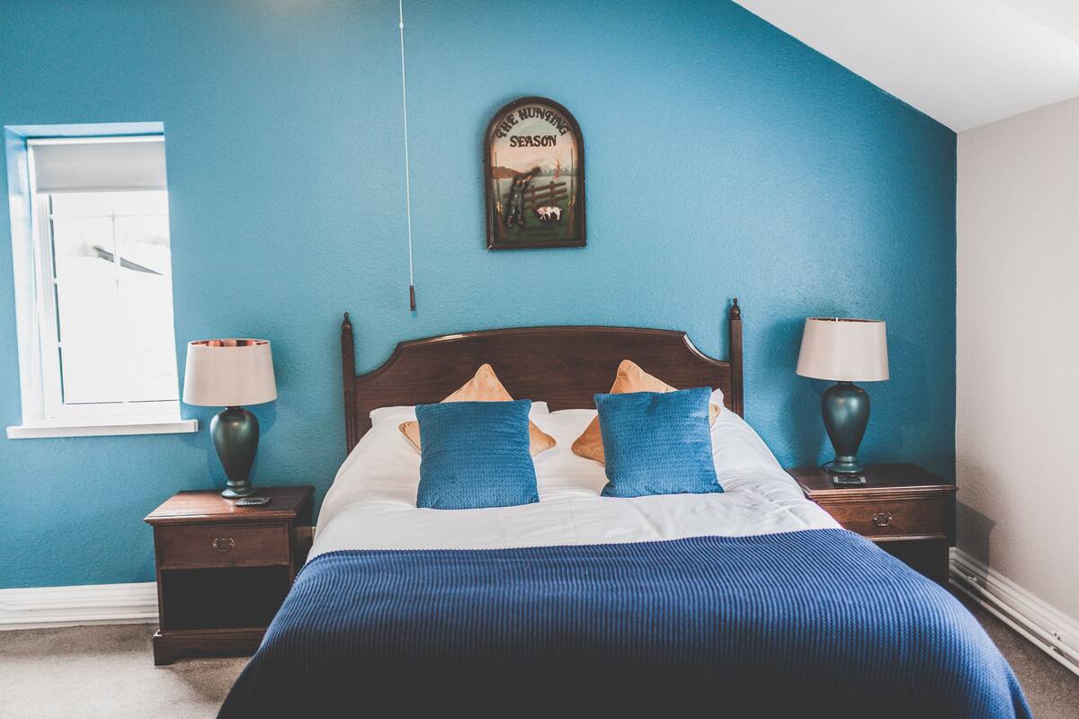 A bedroom with a bed, two nightstands, two lamps, a window, and a wall-mounted picture.