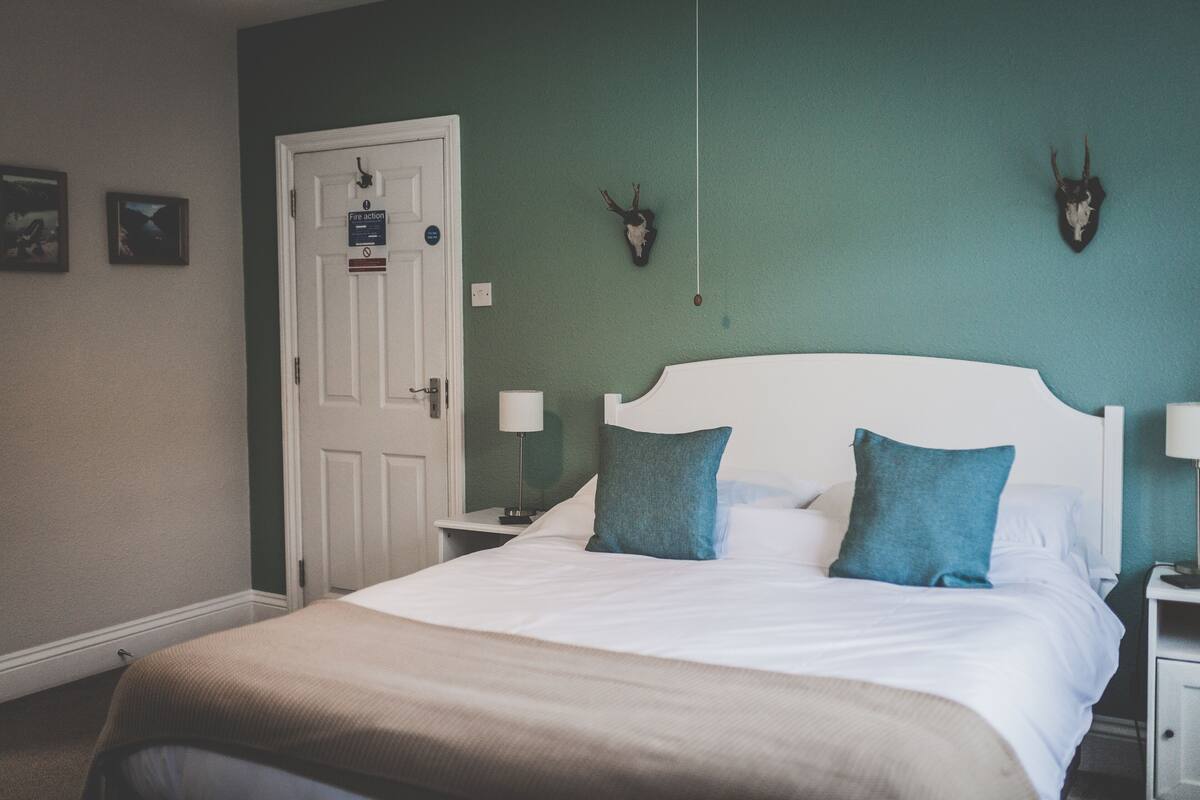 A bedroom with a bed, white headboard, two bedside tables with lamps, a door, and two wall-mounted animal skulls.