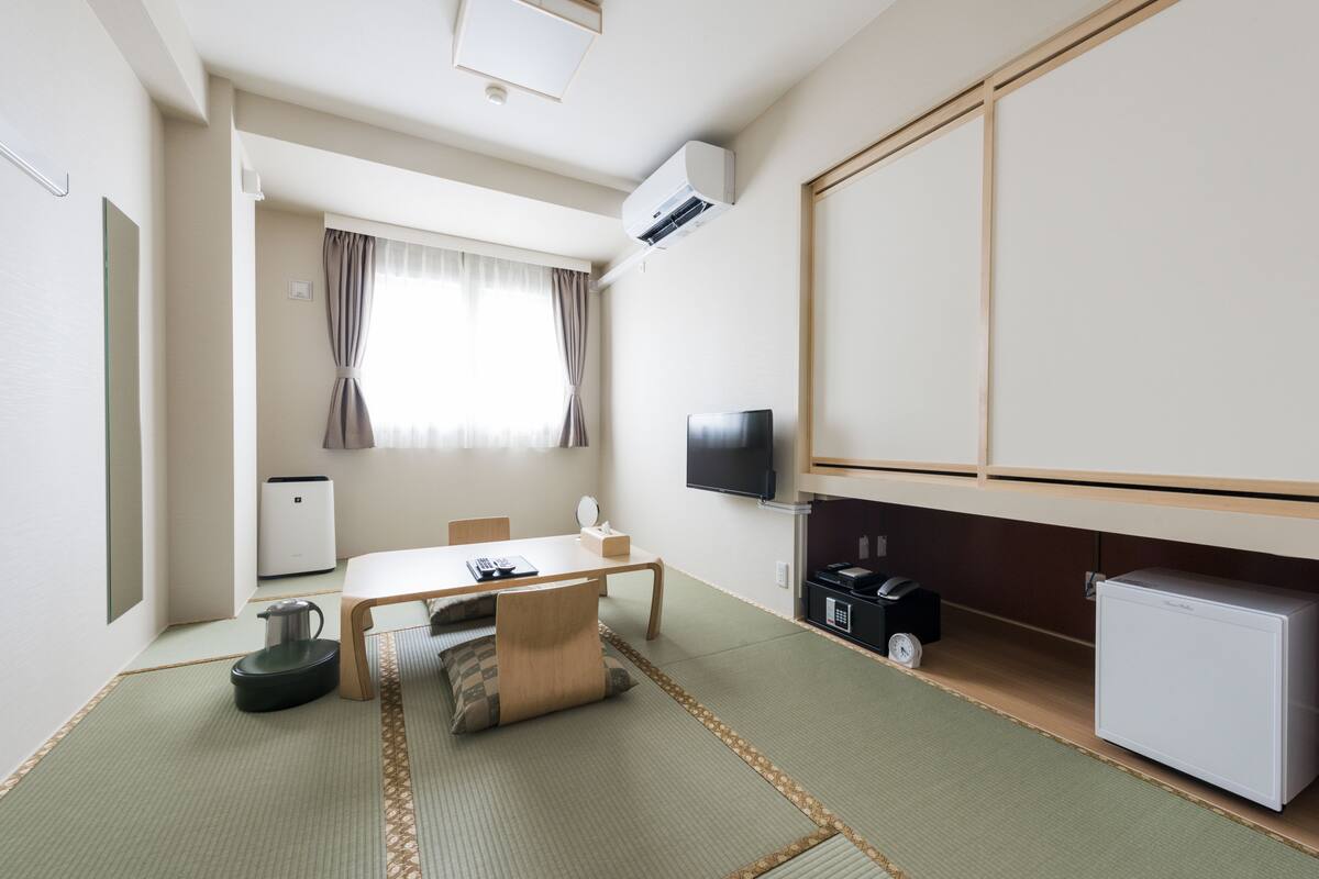 Une chambre d’hôtel compacte dotée d’un sol recouvert de tatamis, d’une petite table avec une chaise, d’un téléviseur, d’un mini-réfrigérateur et d’un radiateur.