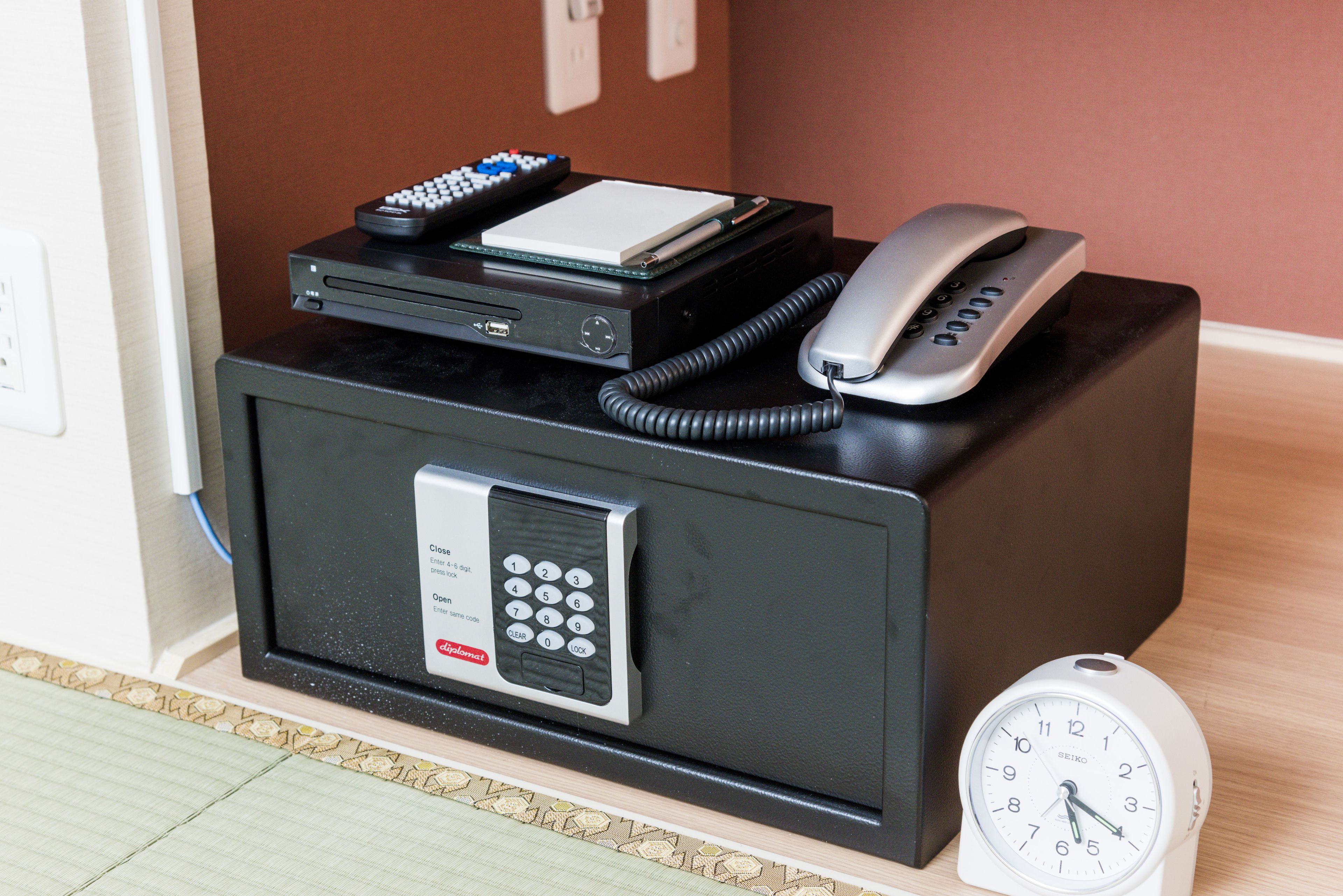 A hotel room with a desk featuring a telephone, a clock, and a small electronic device.