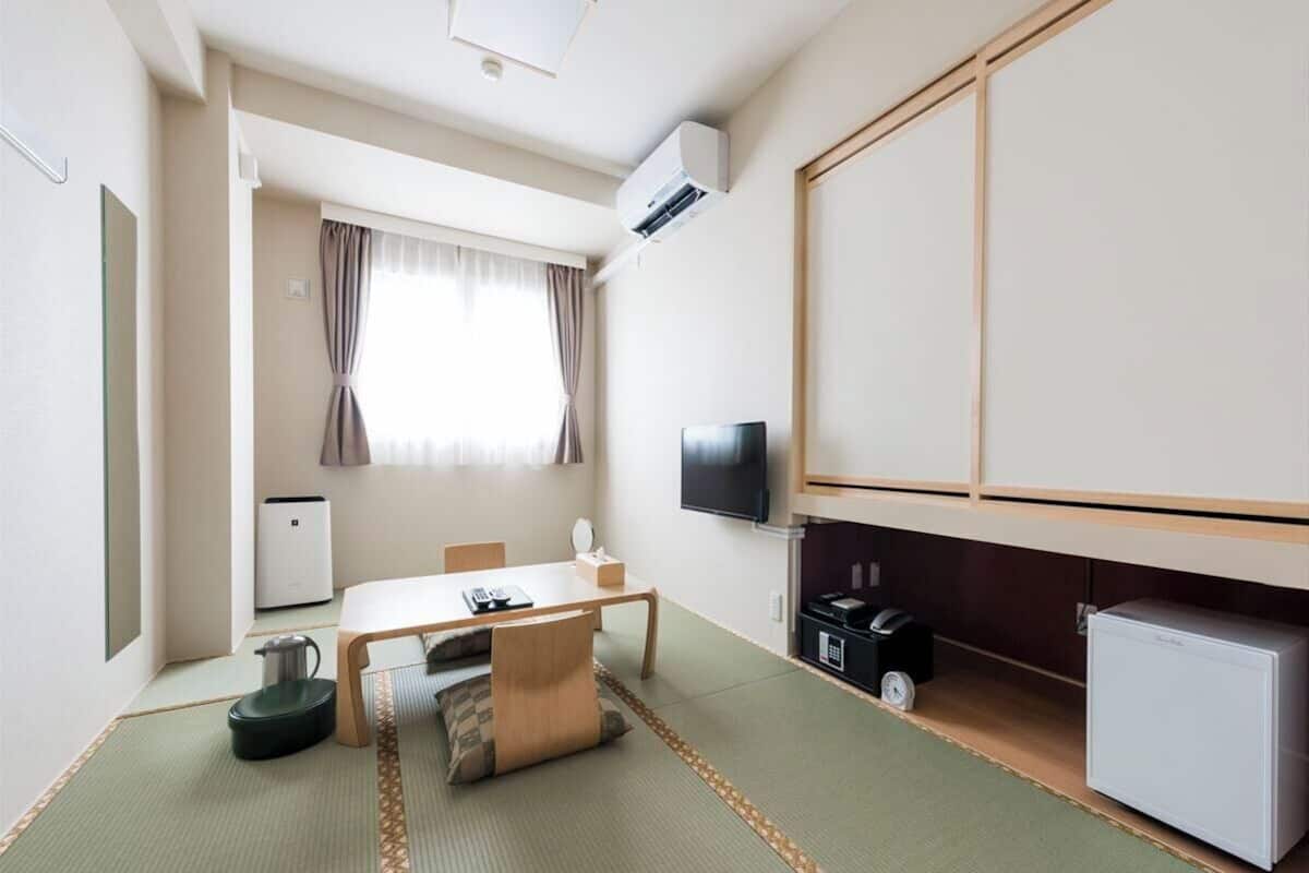 Une chambre d’hôtel avec un sol recouvert de tatamis, une télévision, un petit réfrigérateur et un bureau avec une chaise.