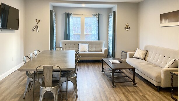 A modern living room with a dining table, two sofas, and a TV mounted on the wall. - Roe Summer Cottage, Suite B (Watkins Glen)