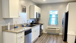 A modern kitchen with white cabinets, a granite countertop, stainless steel appliances, and a window with blue curtains. - Roe Summer Cottage, Suite B (Watkins Glen)