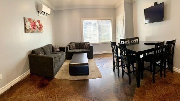 A living room with a sectional sofa, a coffee table, a dining table with chairs, a TV mounted on the wall, and a window with blinds. - Watkins Glen 3-Bedroom Suites Unit 5 (Watkins Glen)