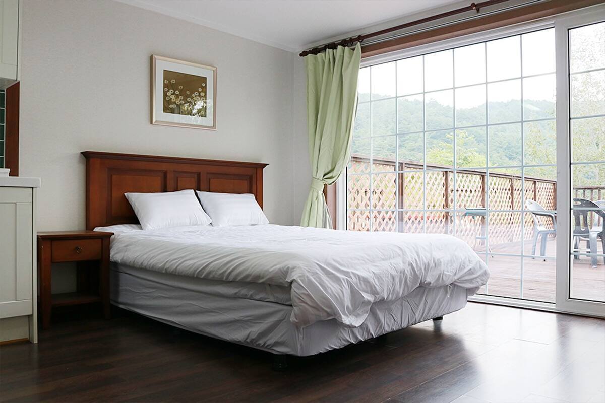 Ein Schlafzimmer mit einem hölzernen Bett, einem Nachttisch, einem gerahmten Bild an der Wand und einem großen Fenster mit Blick auf Grünfläche.