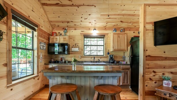 A cabin-style kitchen with wooden walls, a large window, a sink, a refrigerator, and two stools. - God's Country Cabins- Mercy I Close to Town (Fredericksburg)