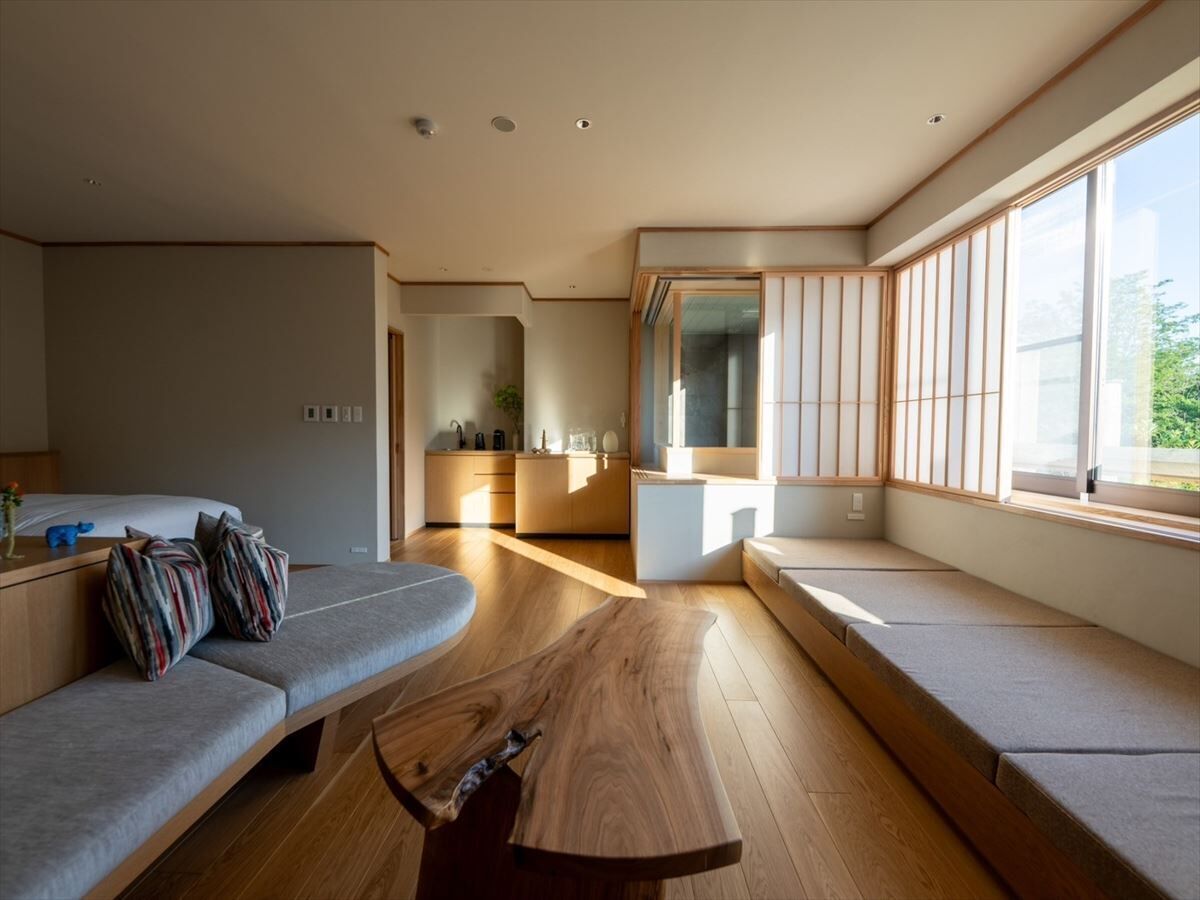 Ein modernes Wohnzimmer mit einer Couch, einem Holztisch und Blick auf eine Küche und Essbereich durch große Fenster.