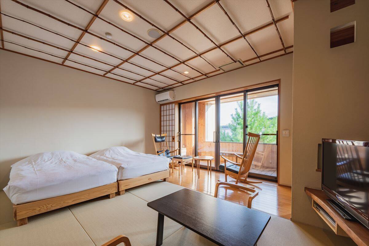Una habitación con dos camas, una mesa, sillas, un televisor y un ventanal corredizo a un espacio exterior.