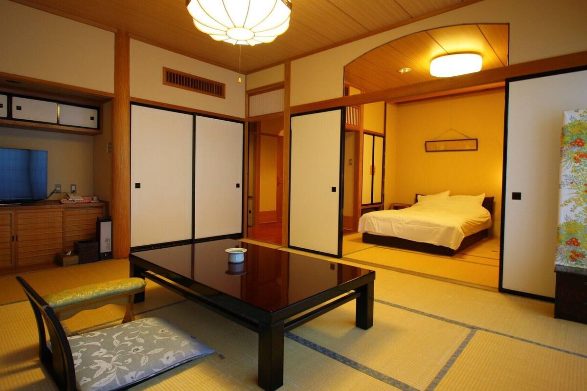 Una habitación japonesa tradicional con suelo de tatami, una mesita baja, una cama con ropa blanca y un televisor sobre un mueble.