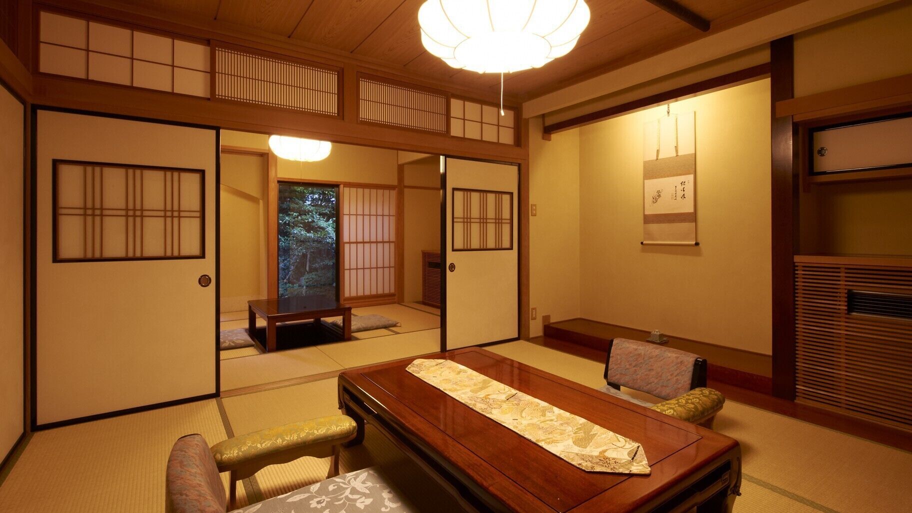 Una habitación japonesa tradicional con suelo de tatami, una mesa baja con un tapete decorativo, puertas correderas y una lámpara en el techo.