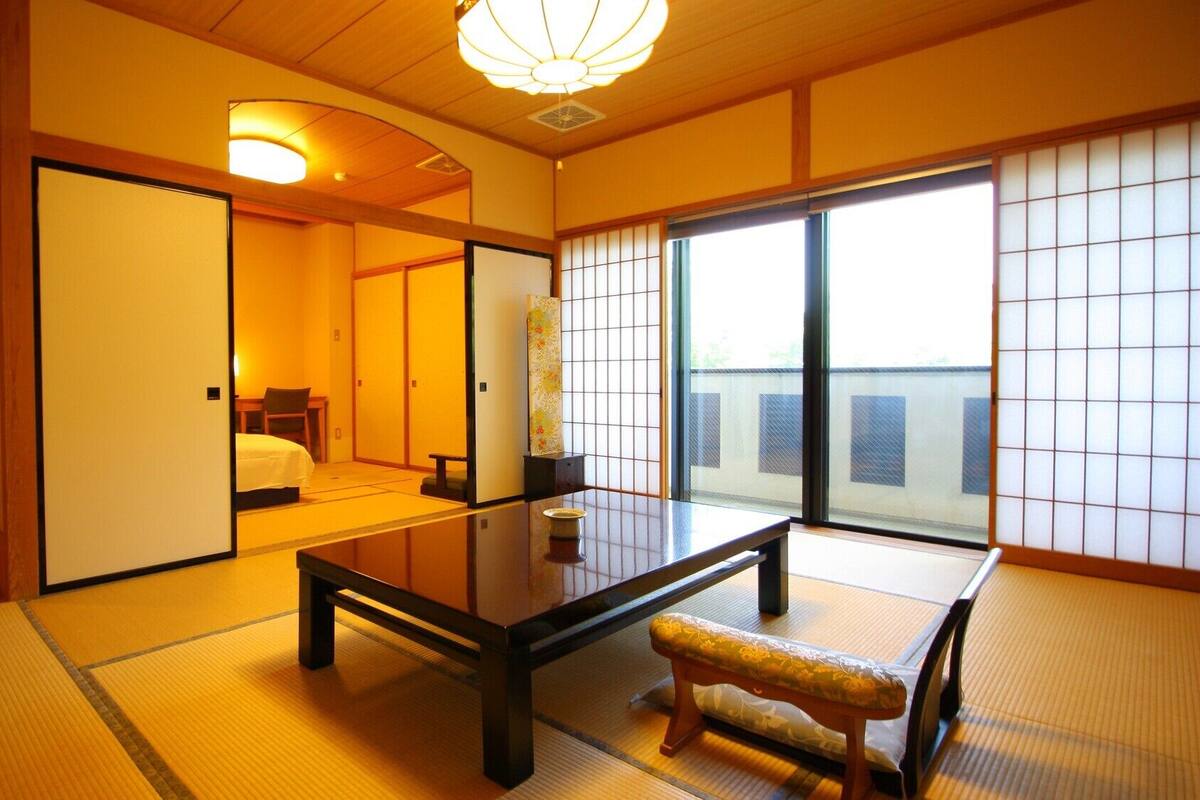 Una habitación tradicional japonesa con suelo de tatami, una mesa baja, un banco, puertas correderas y vistas a través de un celosía shoji.