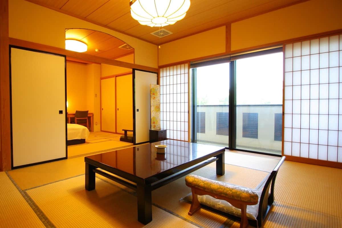 Una habitación tradicional japonesa con suelo de tatami, una mesa baja, un banco, puertas correderas y vistas a través de un celosía shoji.