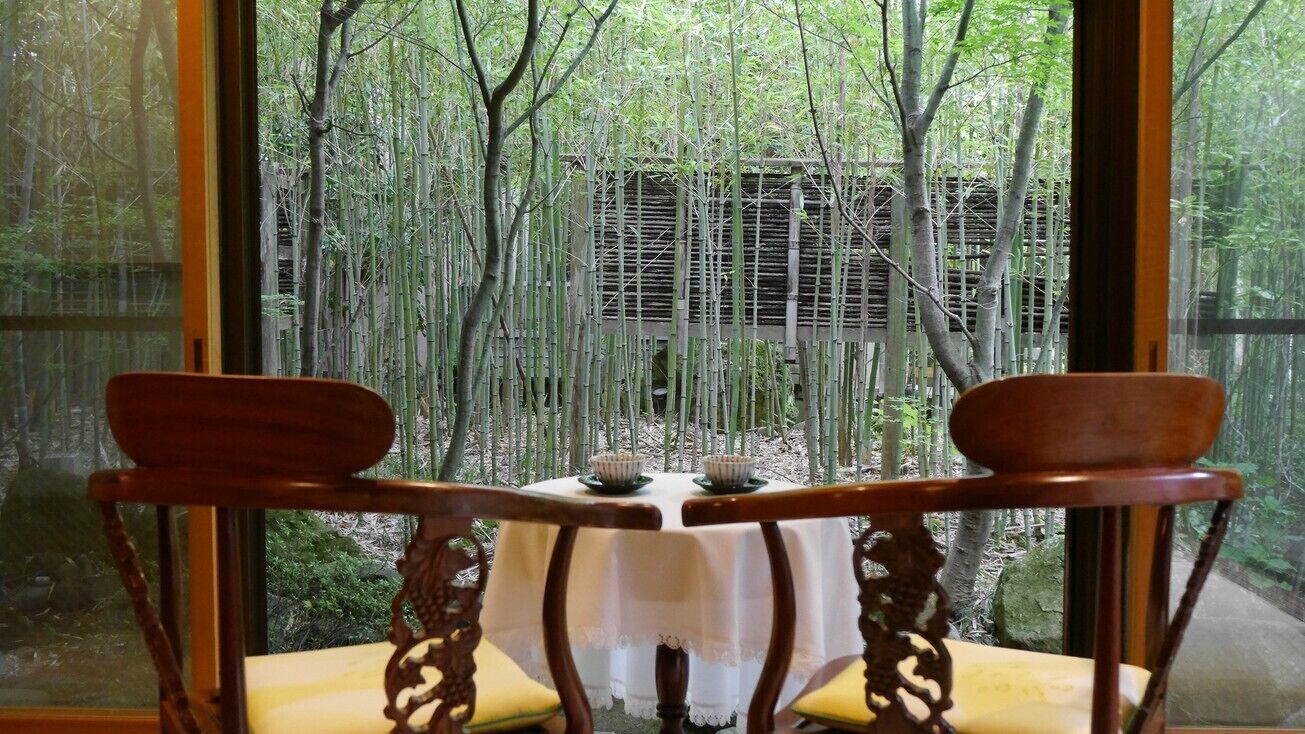 Una mesa con mantel blanco, dos sillas de madera y vistas a un bosque de bambú a través de amplios ventanales.