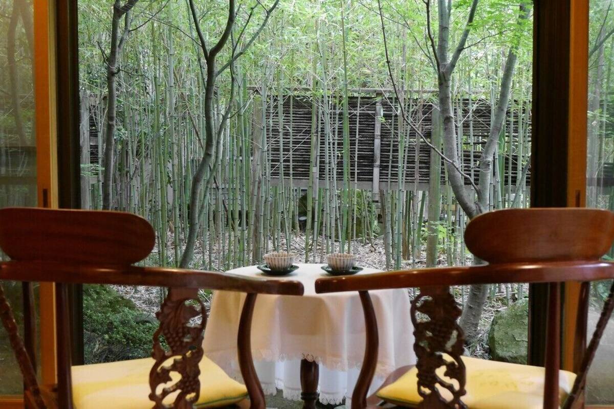 Una mesa con mantel blanco, dos sillas de madera y vistas a un bosque de bambú a través de amplios ventanales.