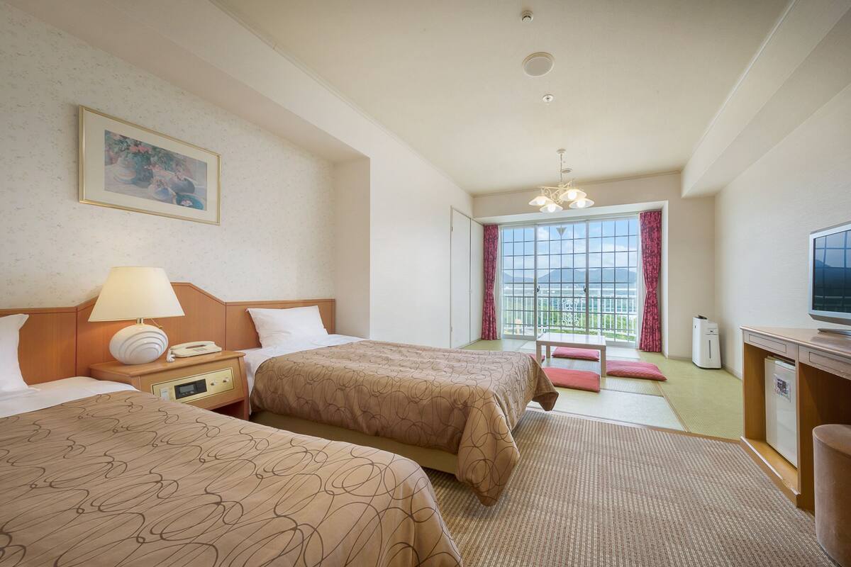 A hotel room with two beds, a desk with a television, a window with a view, and a painting on the wall.