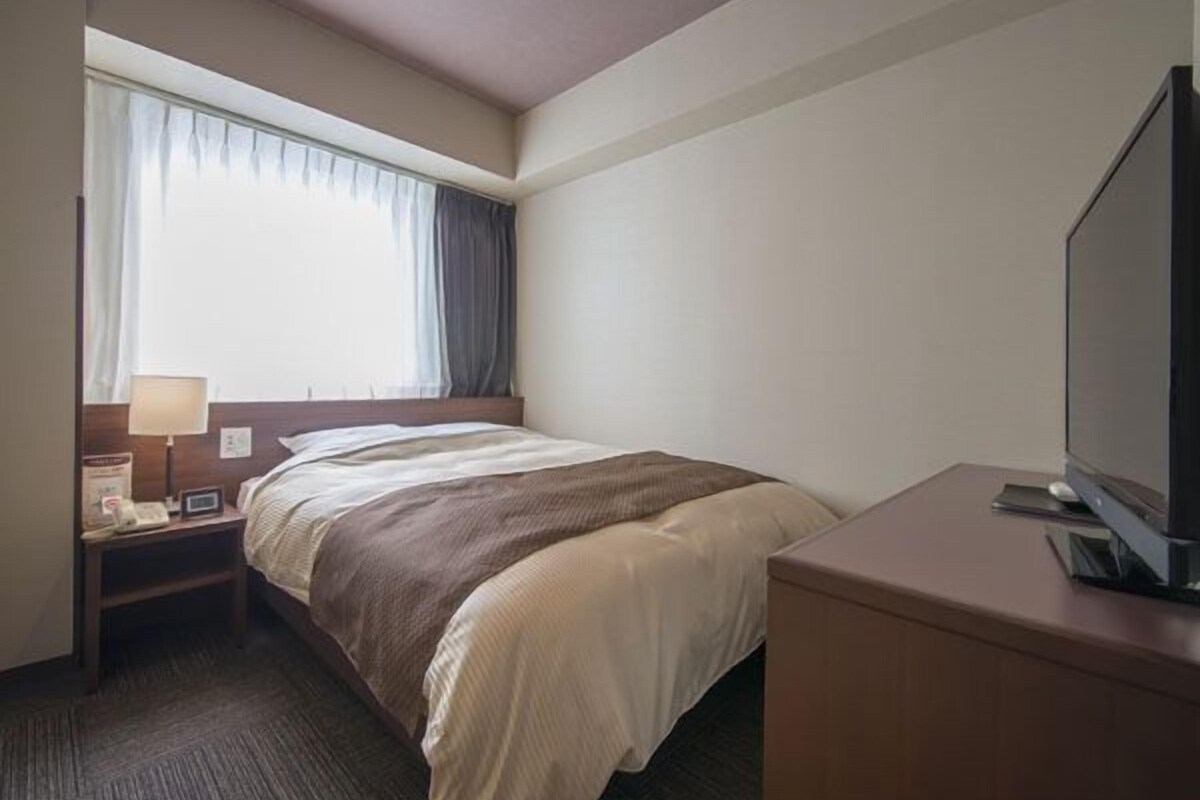 Habitación de hotel con cama, escritorio con televisor de pantalla plana, mesita de noche con lámpara y ventana con cortinas.