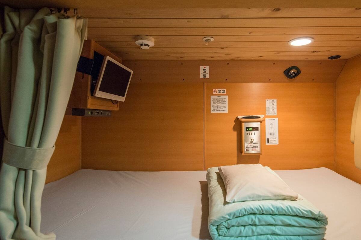 Una habitación pequeña con cama individual, techo de madera, una mesita de noche con lámpara, televisor en la pared y ventana con cortinas.