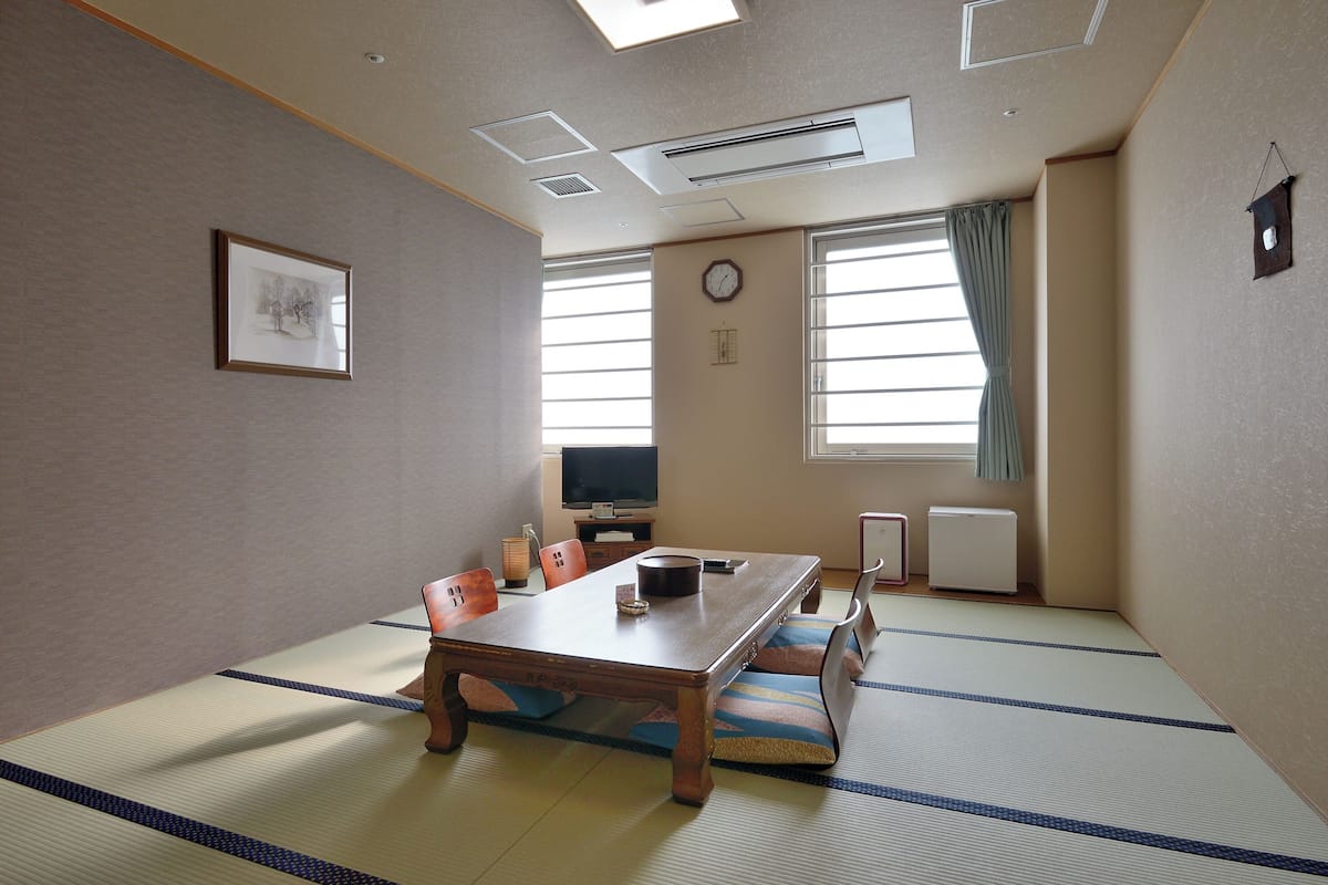 Una habitación japonesa tradicional con suelo de tatami, una mesa baja, sillas, un televisor y un reloj en la pared.