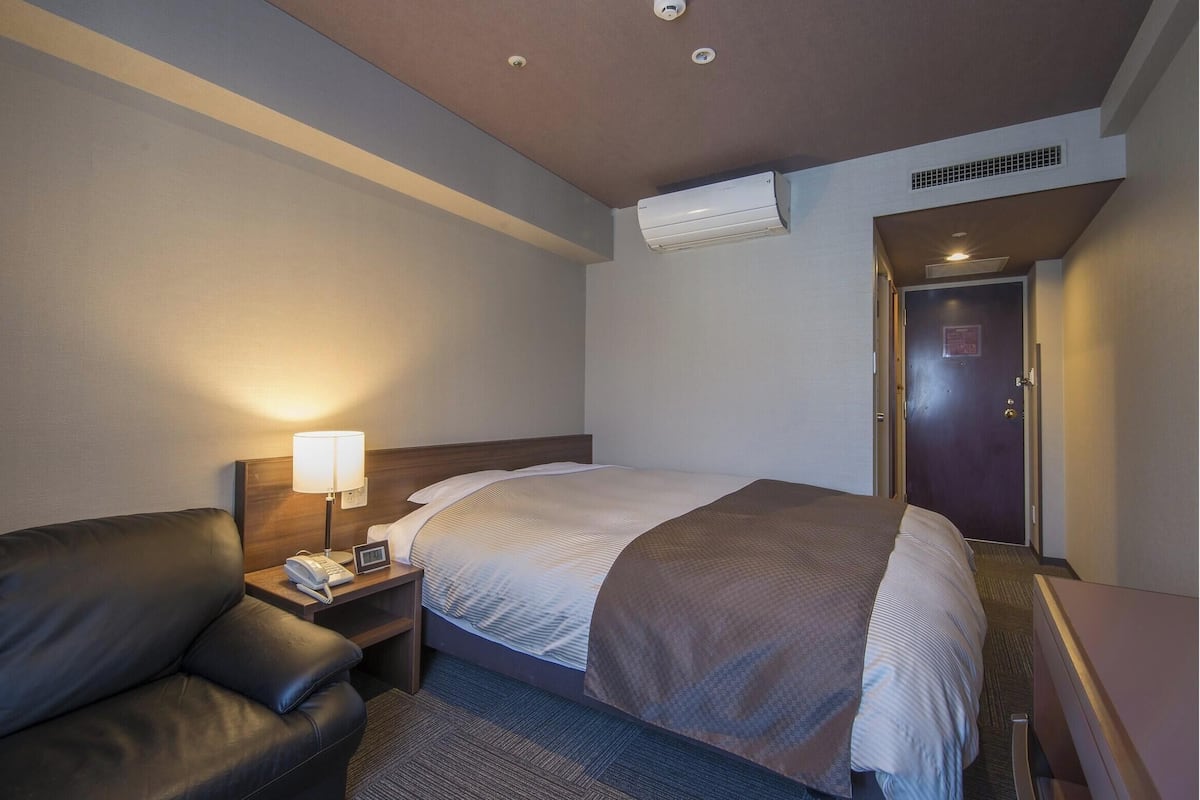 Habitación de hotel con cama, escritorio, silla, lámpara, equipo de aire acondicionado y puerta.