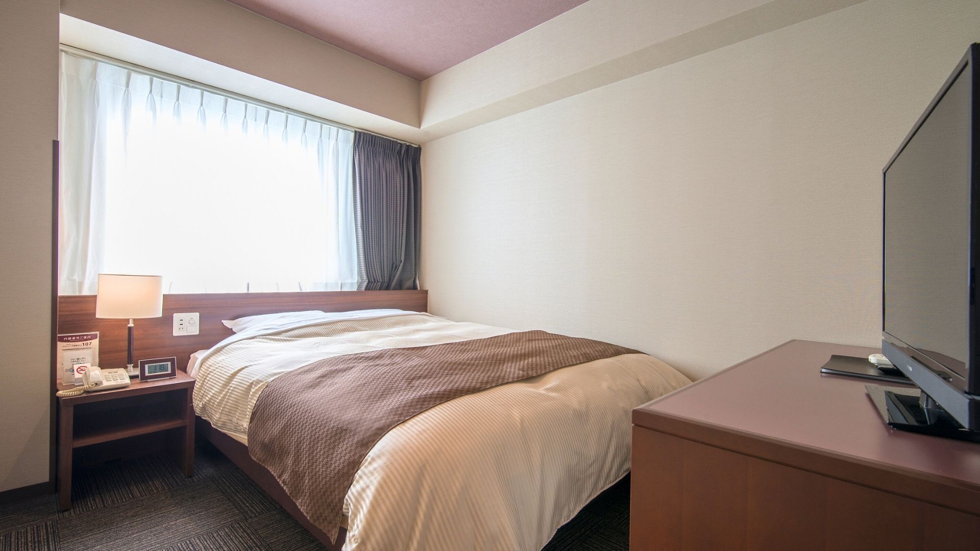 Habitació d'hotel amb llit, taulella de nit, làmpada, rellotge i televisió damunt d'un escriptori.