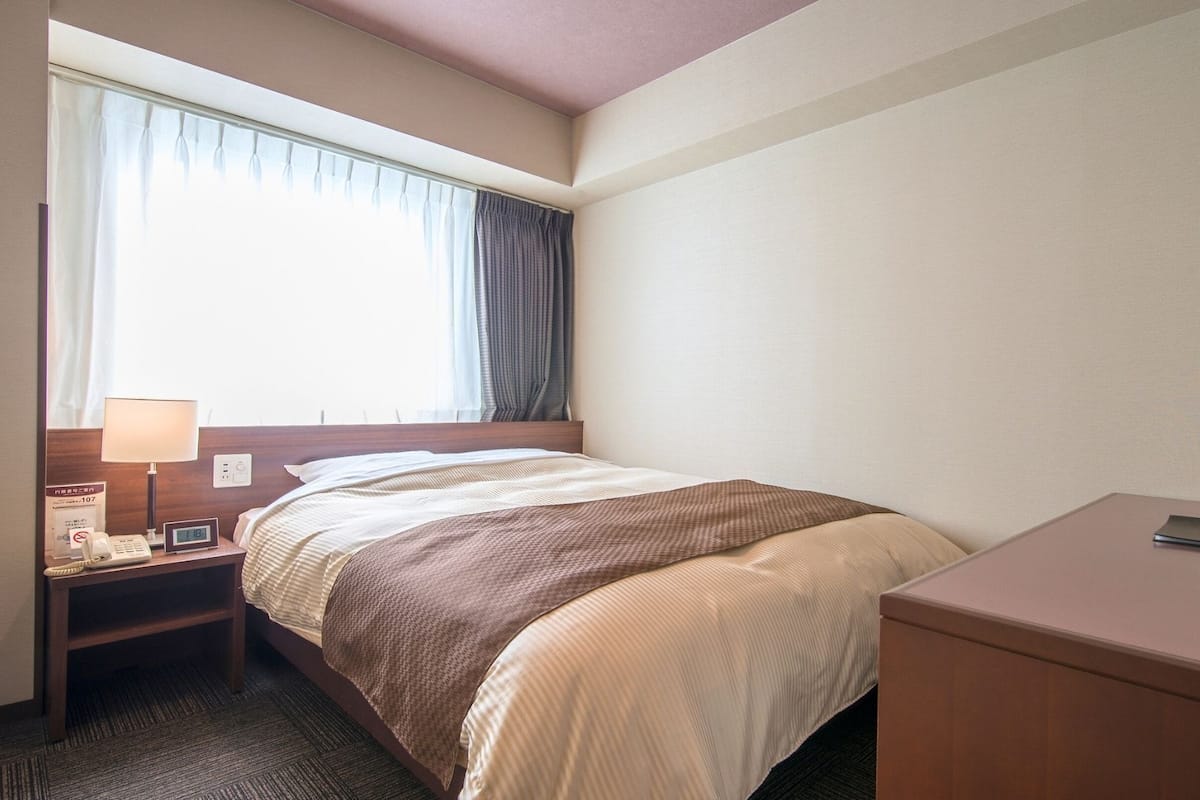 Una habitación de hotel con cama, mesita de noche, lámpara, reloj y televisor sobre un escritorio.
