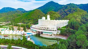 A large, white, multi-story hotel with a unique curved roof, situated by a lake with a bridge connecting to a mainland. - Ooedo Onsen Monogatari Hotel Reoma no Mori (Marugame)