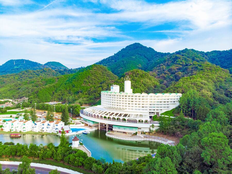 A large, white, multi-story hotel with a unique curved roof, situated by a lake with a bridge connecting to a mainland.